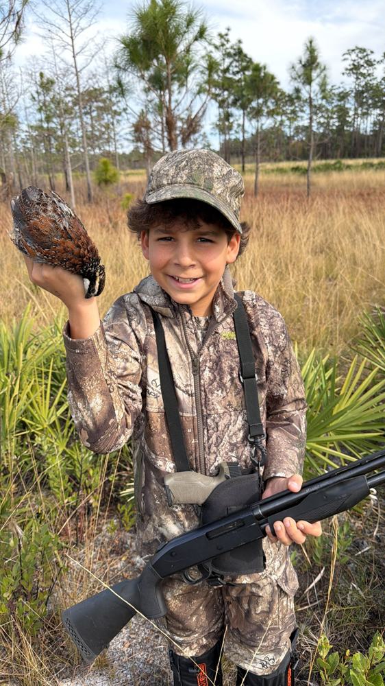 Menino de camuflagem sorrindo no campo, segurando uma codorna na mão e uma espingarda no outro braço