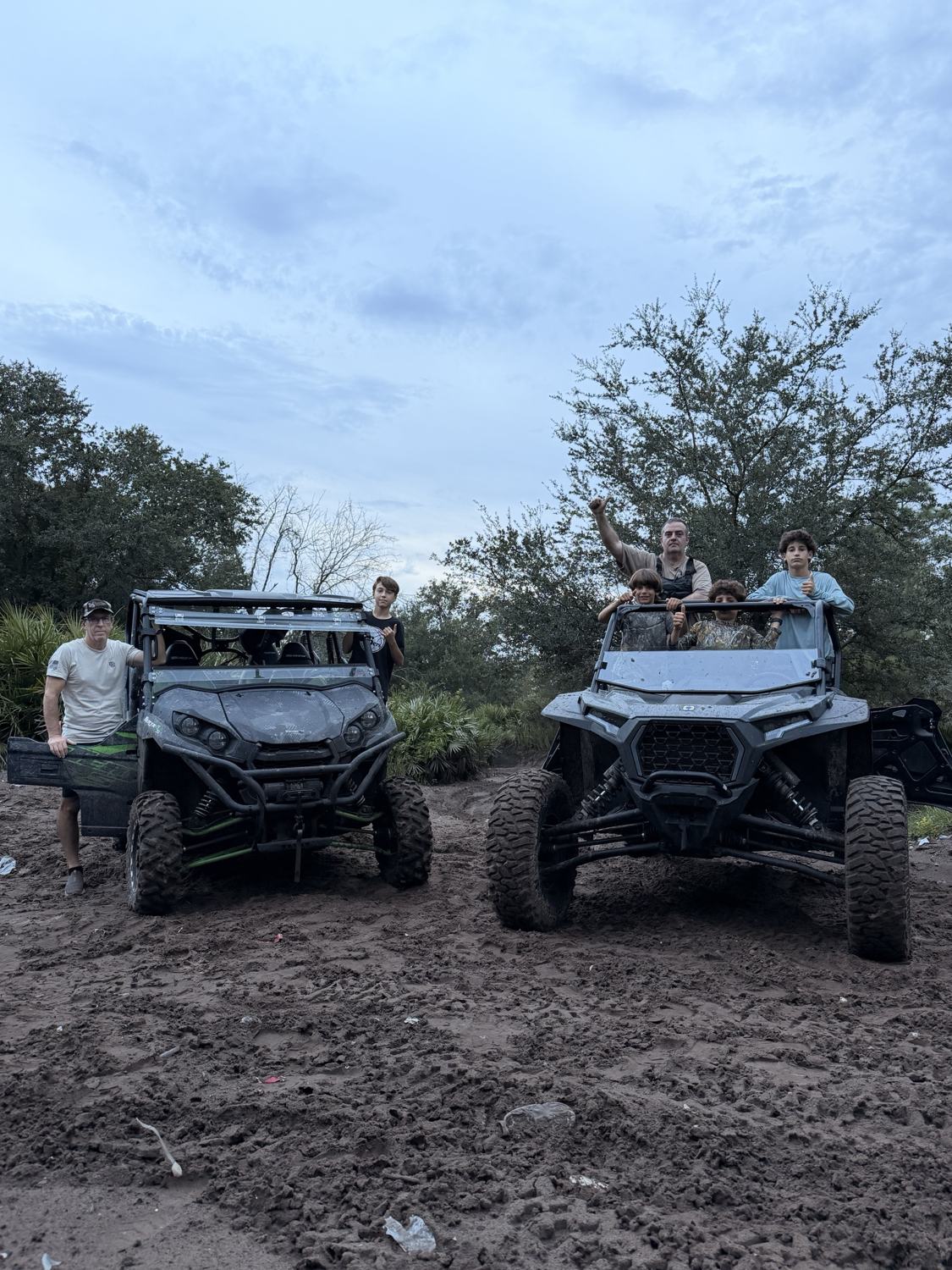Dois UTVs enlameados lado a lado em uma estrada de terra, com a família em cima e em volta