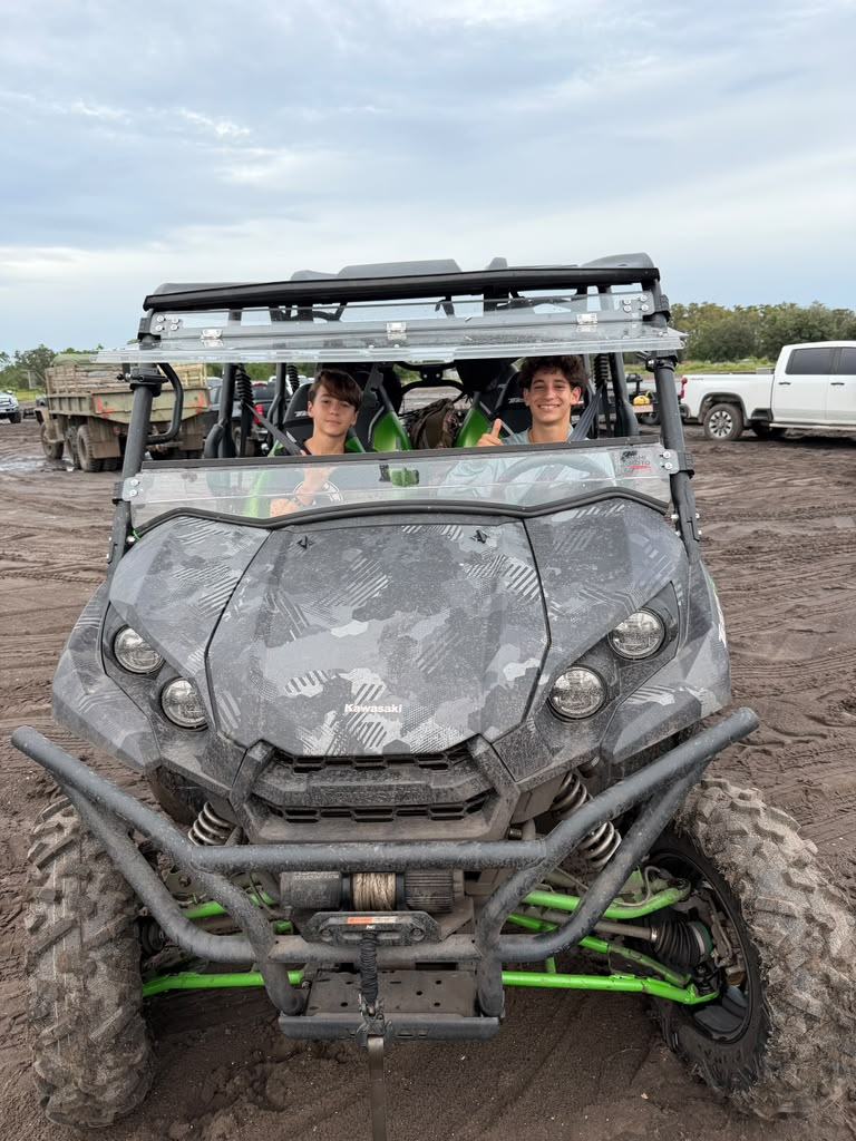 Dois meninos sorrindo dentro de um UTV Kawasaki cinza e verde, parado na lama