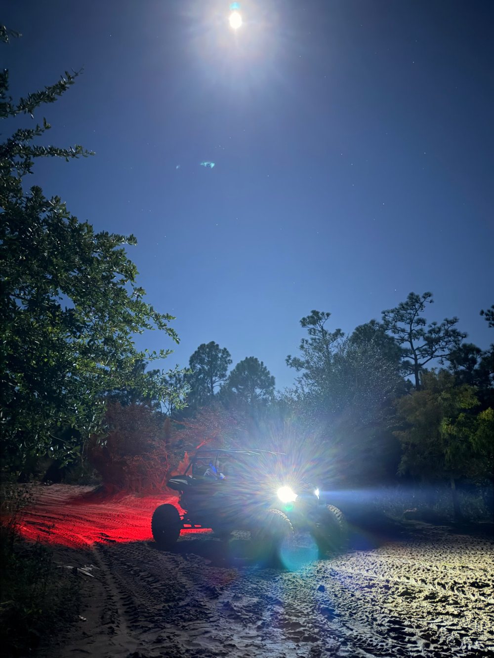 UTV parado numa trilha de areia à noite, com faróis acesos, luz vermelha no chão e a lua no céu