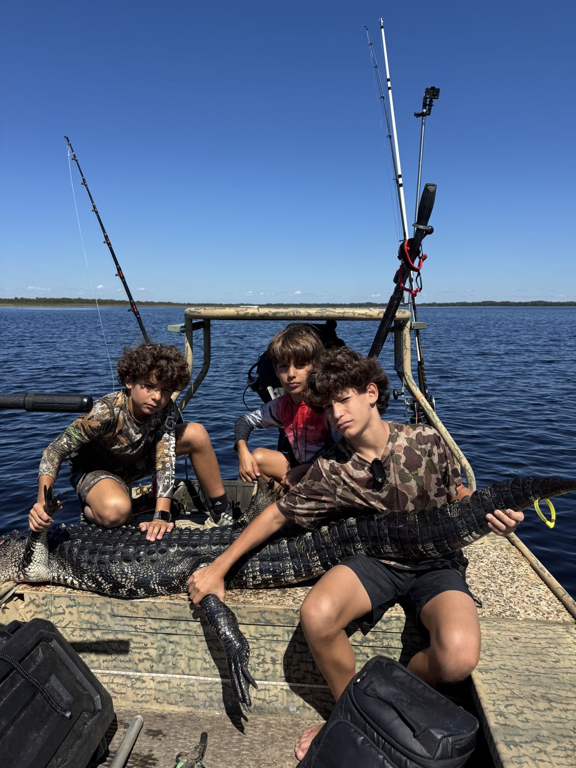 Três meninos em um barco com varas de pesca em um lago, com um jacaré sobre o convés