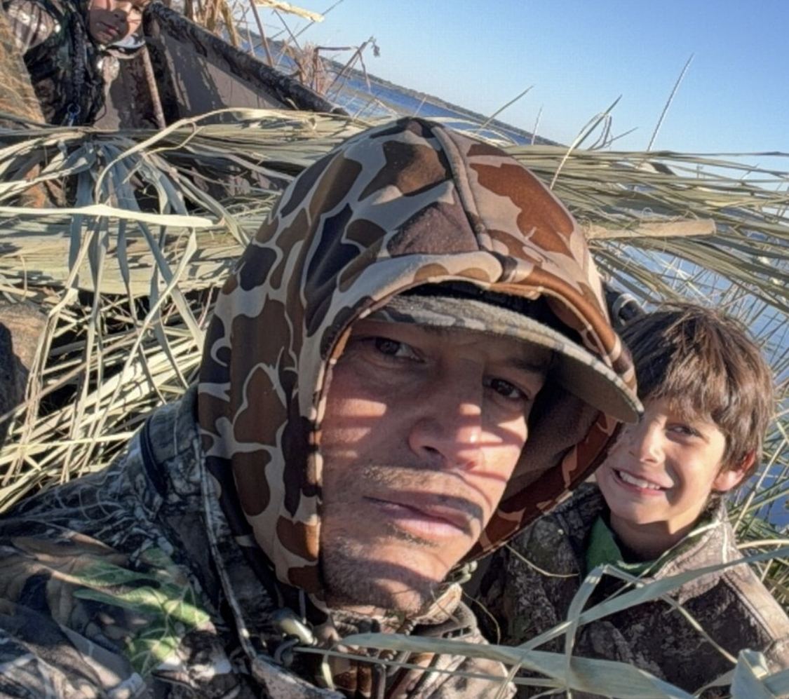 Pai e filho de camuflagem escondidos entre palmeiras à beira de um lago, olhando para a câmera